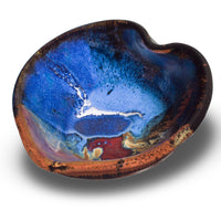 A 5 inch handmade pottery bowl in the shape of a heart.  Beautifully glazed in cobalt blue, toasted orange, and red by Prairie Fire Pottery.  3/4 view.