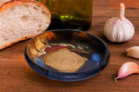 This is a pretty six inch garlic dipping plate in cobalt blue and toasted brown colors . It has a raspy spiral center for breaking up garlic cloves before adding oil and vinegar. Handmade pottery by Prairie Fire Pottery. This is a tabletop view of the product with french bread and olive oil.