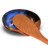 Beautiful milky blue spoon rest accented with toasted brown and red. Hand made in stoneware clay and high-fired to 2400°. It is handmade pottery from Prairie Fire Pottery.  This view is the spoon rest holding a stir spoon.