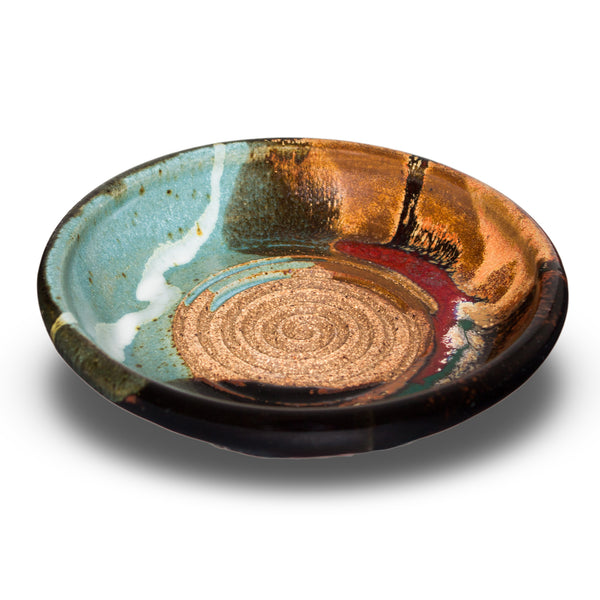 This is a pretty six inch garlic dipping plate in turquoise-brown colors and red. It has a raspy spiral center for breaking up garlic cloves before adding oil and vinegar. Handmade pottery by Prairie Fire Pottery.  This is a 3/4 view of the product.