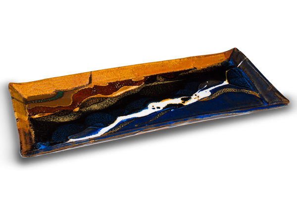 This is high-fired stoneware by Prairie Fire Pottery. It is a 12 1/2 inch rectangular plate in a beautiful blend of cobalt blue, toasted brown, and red. It is hand made and kiln-fired to 2400°. Made in the U.S.A. 3/4 table top view.